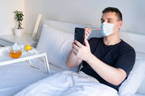 Een man met een verkoudheid in een masker belt de dokter en toont een thermometer. online geneeskunde. - Foto, afbeelding
