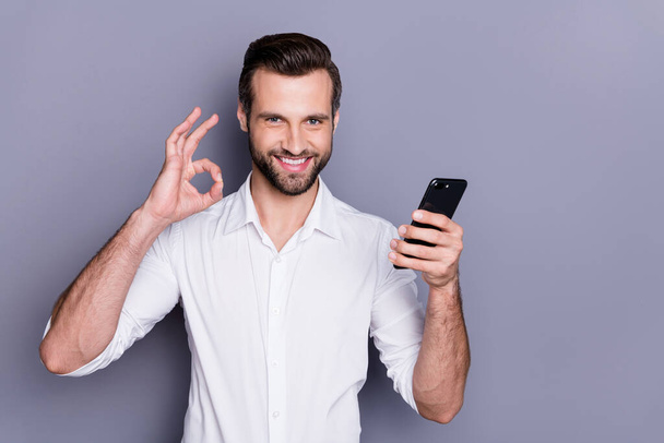 Portrait of positive confident man marketer agent man use smart phone search excellent social network ads approve quality wear white shirt isolated over gray color background - Foto, imagen