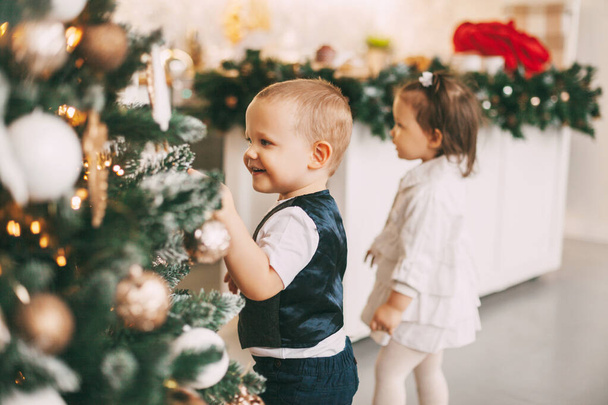 Een grappig jongetje in een pak versiert een kerstboom, zijn zusje staat naast hem. Vakantie, evenement, Kerstmis en Nieuwjaar - Foto, afbeelding