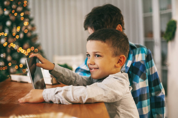3人の男の子は、家族や友人に陽気なクリスマスと幸せな新年を望むためにタブレットを使用しています。子供たちはビデオリンクを介して友人と通信します - 写真・画像