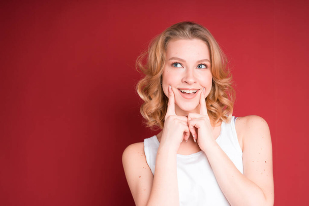 Blond woman with curly hair making smile with her fingers in white tank top isolated on red background. - Photo, Image