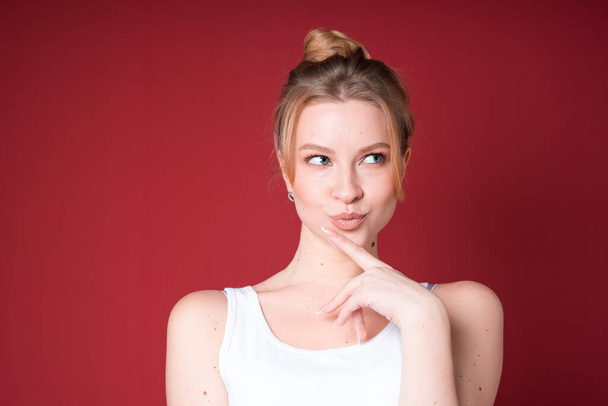Cute blond woman with bun looks dreamy or thoughtful and looking to the side in white tank top isolated on red background. - Foto, imagen