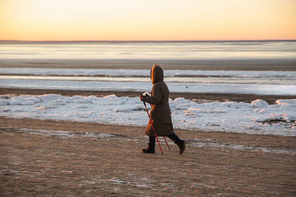 Winter sport in the north - nordic walking. Senior woman hiking in winter sea beach. Active people outdoors. Scenic peaceful sunset landscape - Fotó, kép