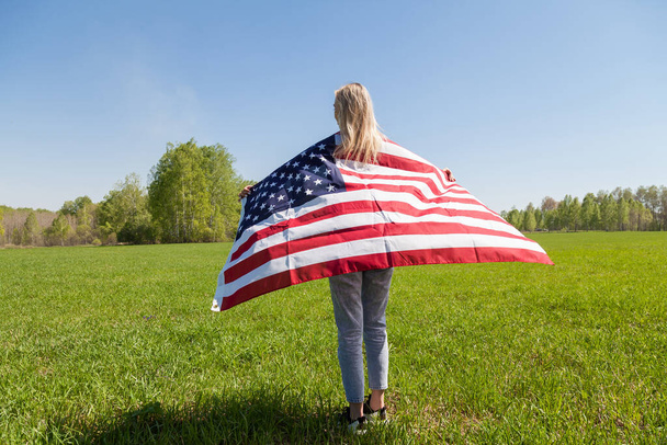 Young woman with blond hair and a flag of the United States of America on her back in blue jeans and sneakers in a field against a background of green trees and grass. Rally in USA. - Fotó, kép