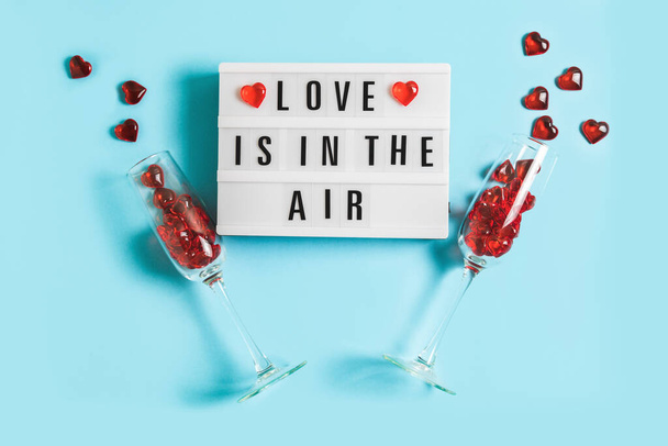 Festive background with glowing lightbox with text Love is in the Air and champagne glasses with red hearts on blue background for Valentine's Day. Flatlay for 14th February. Top view - Foto, Imagem