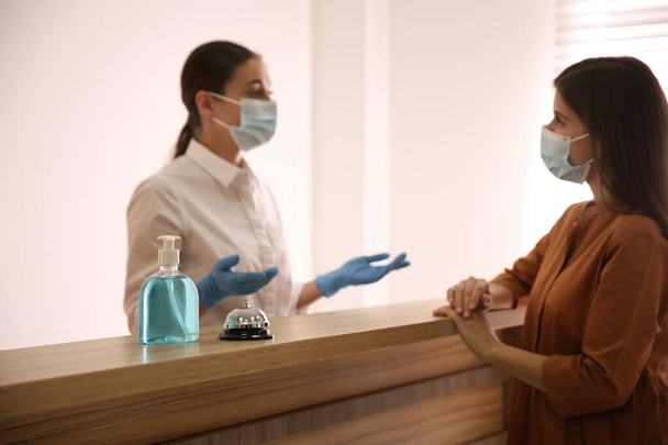 Receptionist with client at countertop in hotel, focus on dispenser bottle of antiseptic gel and service bell - 写真・画像