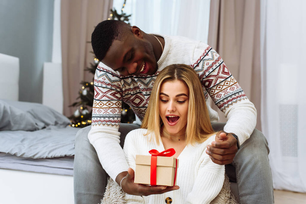Cozy moments in winter holidays at decorated tree. Seasonal greetings concept. A young woman is surprised by a gift that a boyfriend gives her - Photo, Image
