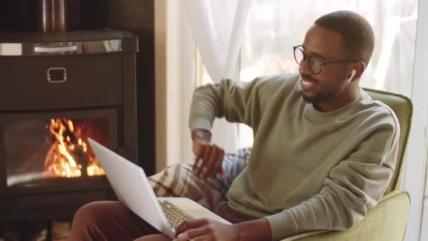 Junger positiver afroamerikanischer Mann mit drahtlosen Kopfhörern lächelt, winkt und plaudert auf einem Laptop, während er im Ferienhaus am Kamin sitzt - Filmmaterial, Video