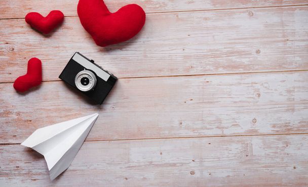 White origami paper airplane, red stuffed plush toy hearts, and black with silver old retro film camera. Wooden flat lay background for Valentines day holiday on February 14. - Fotó, kép