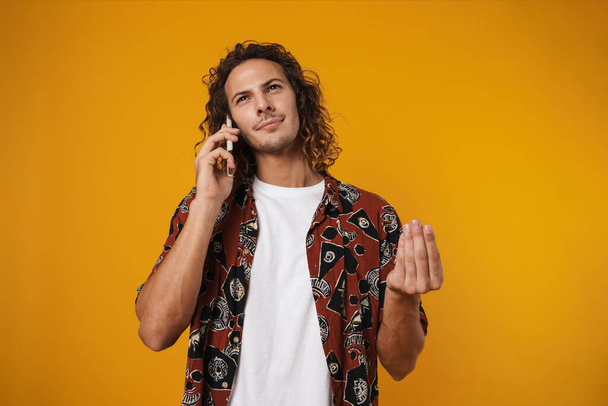 Image of thinking young man talking mobile phone and gesturing isolated over yellow background - Fotó, kép