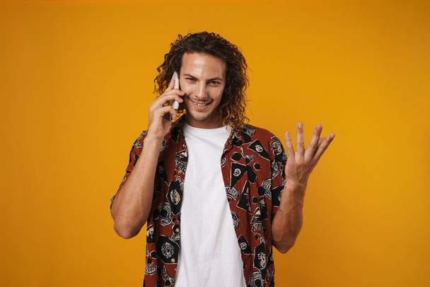 Image of joyful young man talking mobile phone and gesturing isolated over yellow background - 写真・画像