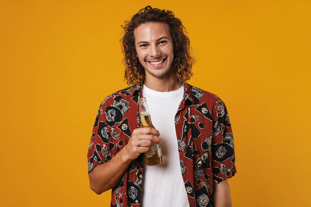Image of cheerful young man smiling and drinking bear isolated over yellow background - Foto, afbeelding