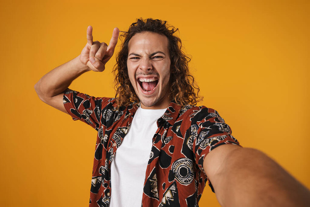 Image of excited young man taking selfie photo and showing horn gesture isolated over yellow background - Foto, Bild