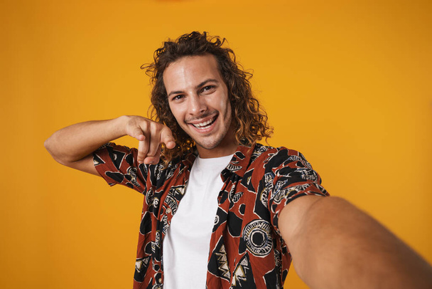 Image of excited young man taking selfie photo and showing horn gesture isolated over yellow background - Foto, immagini