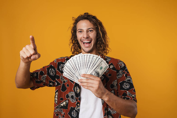 Image of excited young handsome man holding dollars and pointing finger at camera isolated over yellow background - Photo, image
