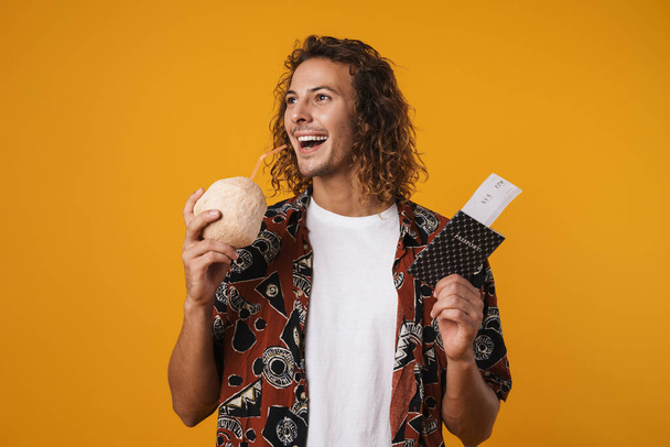 Photo of happy guy drinking coconut water while holding passport with tickets isolated over yellow background - Fotoğraf, Görsel