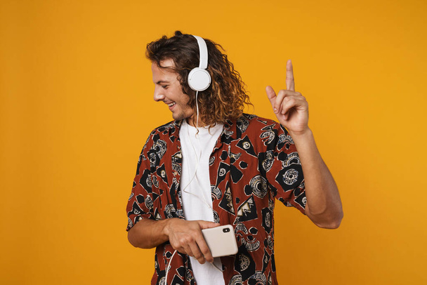 Image of joyful young man in headphones using mobile phone and pointing finger upward isolated over yellow background - Photo, Image