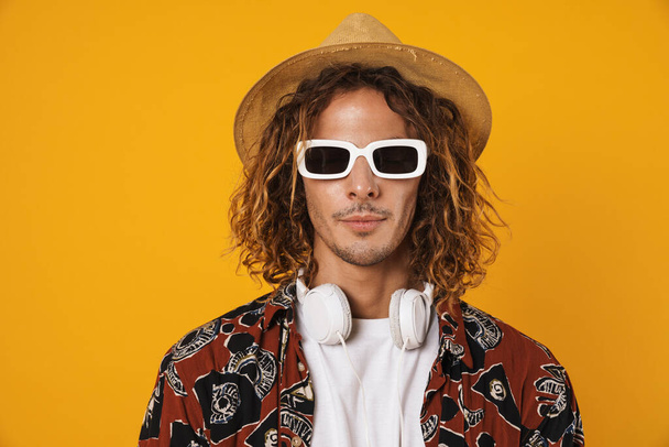 Image of handsome young man in sunglasses and hat posing at camera isolated over yellow background - Fotó, kép