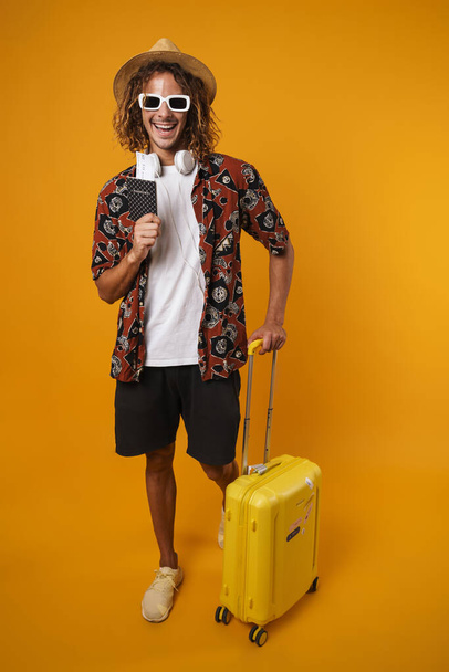 Image of cheerful young man in sunglasses posing with passport and suitcase isolated over yellow background - 写真・画像