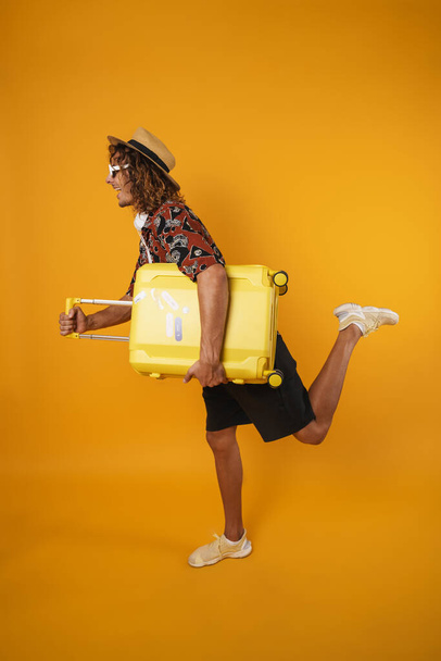 Image of excited young man in hut making fun with suitcase isolated over yellow wall - Fotoğraf, Görsel