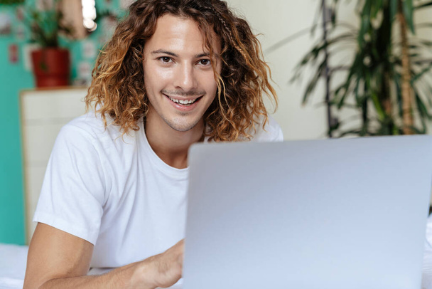 Photo of young positive curly man indoors at home using laptop computer - Φωτογραφία, εικόνα
