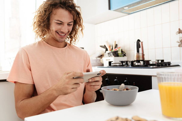 Caucasian smiling guy playing online game on cellphone while having breakfast at kitchen - Fotoğraf, Görsel