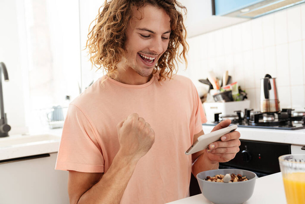 Caucasian excited guy making winner gesture while playing online game on cellphone at kitchen - Valokuva, kuva