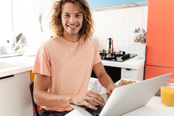 Image of positive young handsome curly man indoors at home using laptop computer - Фото, зображення