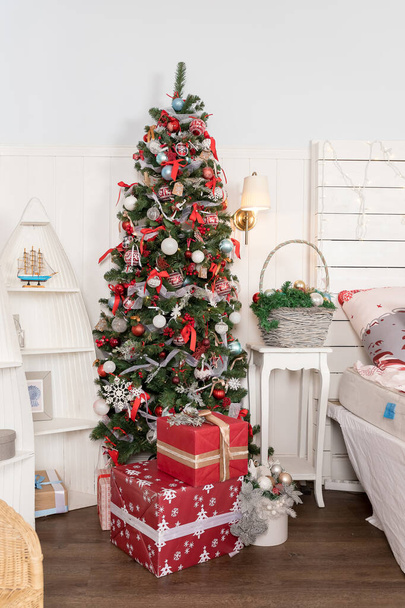 新年の装飾された家のインテリアの木の下で美しいクリスマスプレゼント。クリスマスのお祝い、多色のボールで休日のシーン、装飾的な輝きの弓と木の花に飾られた部屋. - 写真・画像