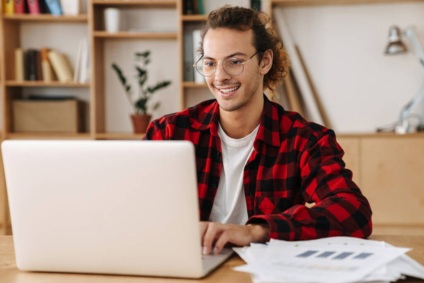 Handsome smiling guy in eyeglasses working with laptop while sitting at office - Фото, изображение