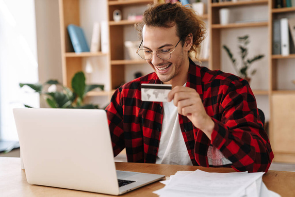 Handsome smiling guy holding credit card and working with laptop at office - Photo, Image