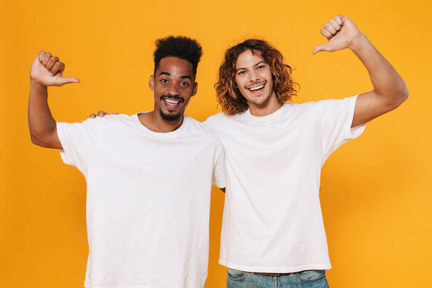 Happy multicultural two guys pointing fingers at themselves and hugging isolated over yellow background - Foto, immagini