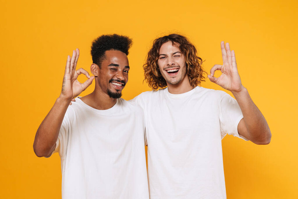Happy multicultural two guys showing ok sign and winking while hugging isolated over yellow background - Valokuva, kuva