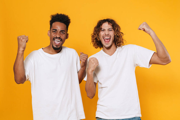 Excited multicultural two guys screaming and making winner gesture isolated over yellow background - Fotoğraf, Görsel