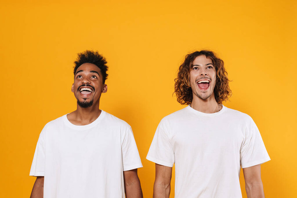 Delighted multicultural two guys exclaiming and looking upward isolated over yellow background - Foto, imagen