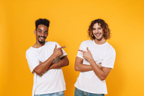 Happy multicultural two guys smiling and pointing fingers at each other isolated over yellow background - Photo, image