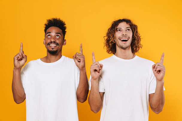 Delighted multicultural two guys looking and pointing fingers upward isolated over yellow background - Fotoğraf, Görsel
