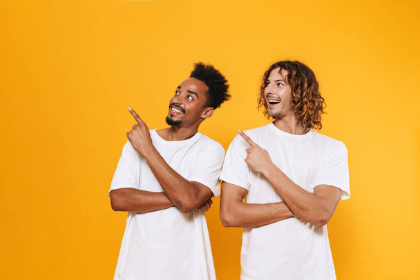 Delighted multicultural two guys looking and pointing fingers aside isolated over yellow background - Fotoğraf, Görsel