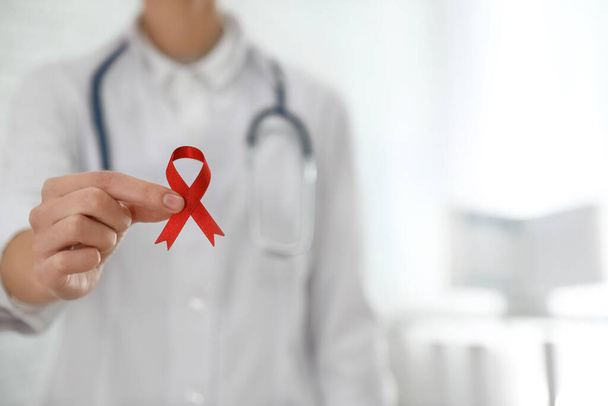 Doctor holding red ribbon on blurred background, closeup with space for text. AIDS awareness month - Φωτογραφία, εικόνα