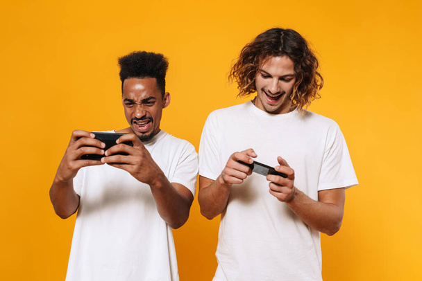 Excited multicultural two guys playing online games on smartphones isolated over yellow background - Valokuva, kuva