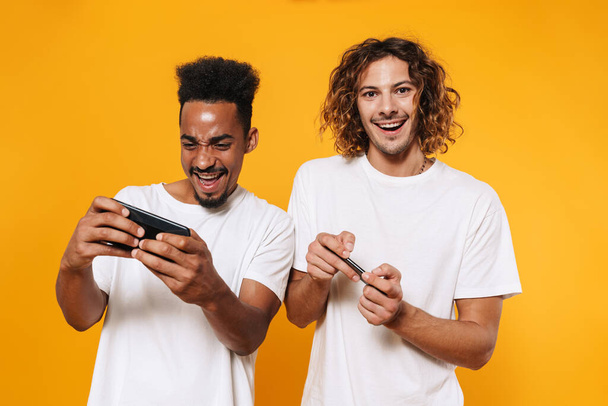 Excited multicultural two guys playing online games on smartphones isolated over yellow background - Photo, image