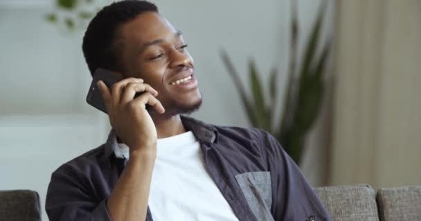 Πορτρέτο του χαμογελαστού μαύρου άντρα χαρούμενο αρσενικό πρόσωπο Αφροαμερικανός που κάθεται ξεκουράζεται χαλαρώνει στο σπίτι στον καναπέ μιλάει στο smartphone επικοινωνεί, ελεύθερος επαγγελματίας λειτουργεί εξ αποστάσεως στο τηλέφωνο να κάνει τις επιχειρήσεις σε απευθείας σύνδεση - Πλάνα, βίντεο