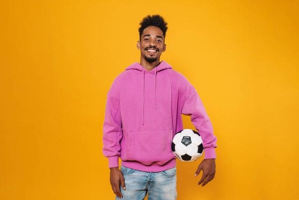 Heureux homme afro-américain souriant tout en posant avec le football isolé sur fond jaune - Photo, image
