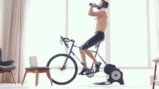 Увлажняйся. Молодой спортсмен, велосипедист в наушниках, пьющий воду или изотонический напиток во время тренировки на стационарном велосипеде дома. Велоспорт в помещении, спиннинг - Кадры, видео