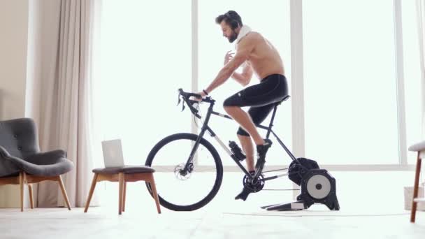 自宅で働いています。若いスポーツマンのサイクリストは、自宅で静止運動用自転車のトレーニング中にタオルで顔から汗を拭くことに集中しました。屋内サイクリング、回転 - 映像、動画