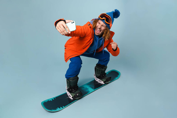 Handsome delighted sportsman taking selfie on cellphone while snowboarding isolated over blue background - Фото, зображення
