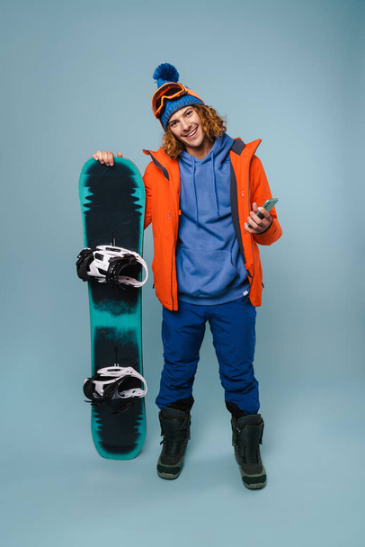 Handsome happy sportsman posing with cellphone and snowboard isolated over blue background - Valokuva, kuva