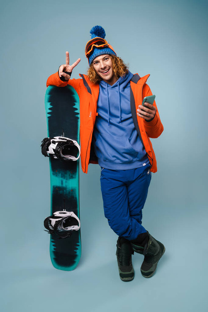Przystojny szczęśliwy sportowiec pozowanie z telefonu komórkowego i snowboardu izolowane na niebieskim tle - Zdjęcie, obraz