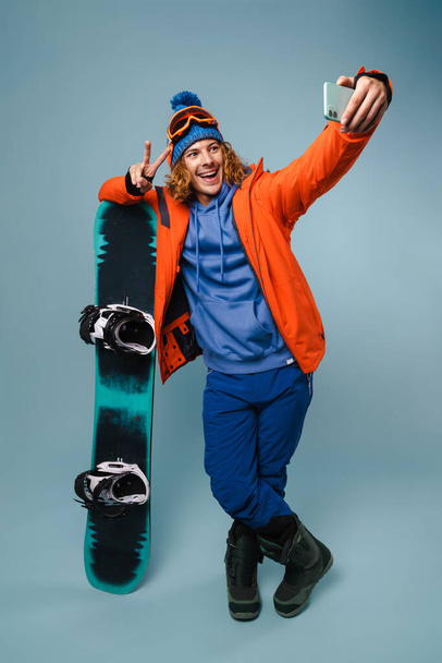 Handsome happy sportsman taking selfie with snowboard on cellphone isolated over blue background - 写真・画像