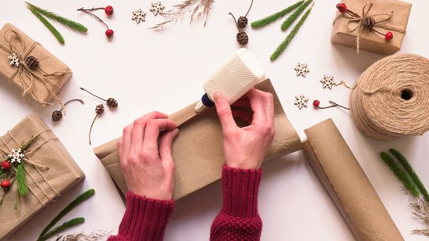 Stap 3. Stap voor stap instructie om milieuvriendelijke en nul afval kerstcadeau verpakking. Vrouw wikkelt geschenk in bruin papier en verzegelt randen met lijm, bovenaanzicht, plat lay - Foto, afbeelding